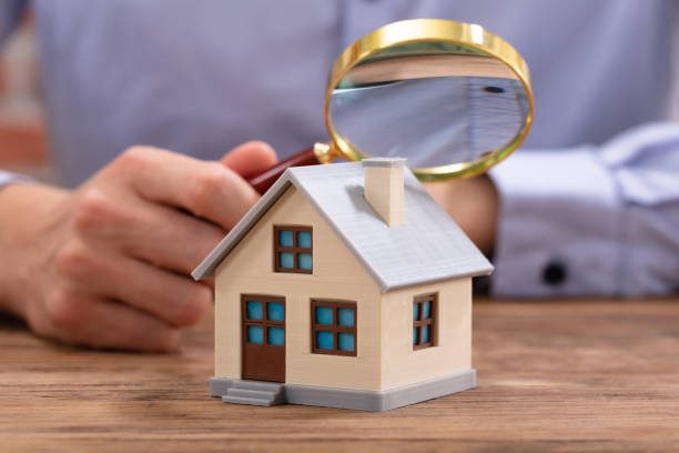 Close-up Of A Businessman's Hand Holding Magnifying Glass Over House Model Over Desk